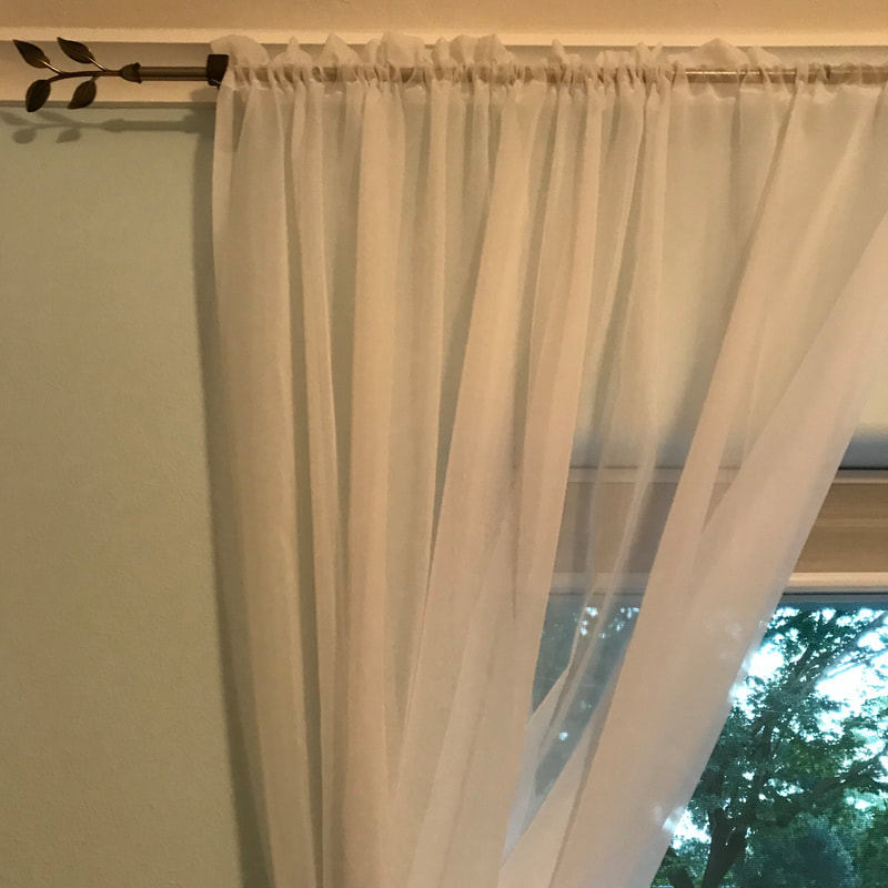 White filmy see-through curtains hanging across the corner of a window.  Trees in the background.  The curtain is held up by a bronze curtain rod with four decorative leaves at the end.  White crown moulding.  Pale blue wall paint.  