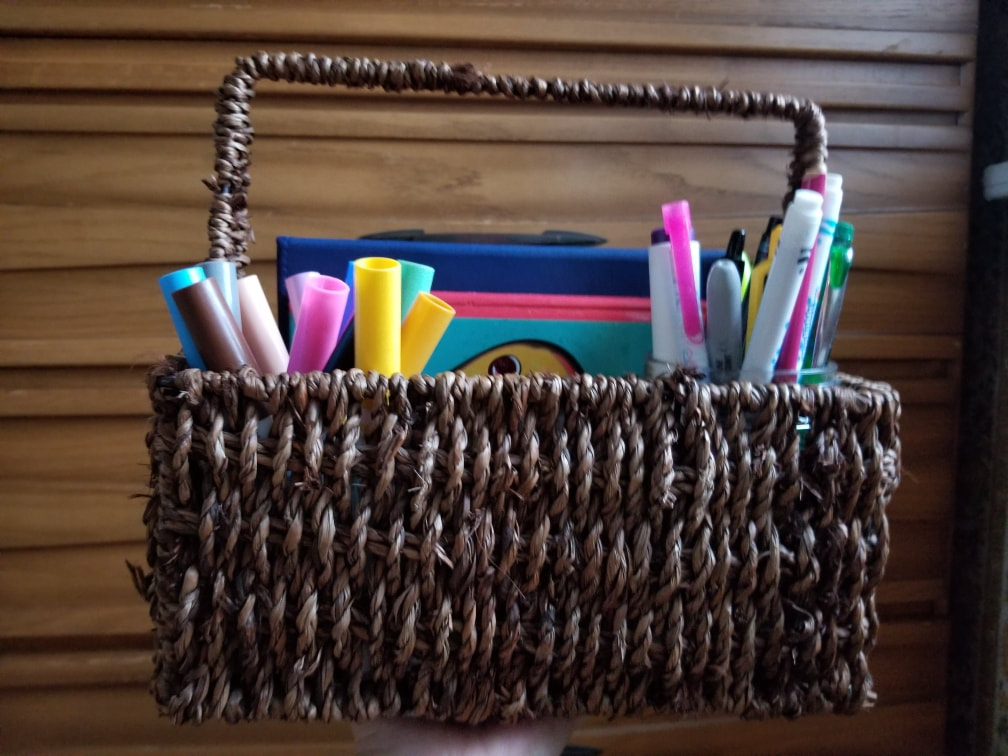 Dark wood slatted background with a dark wood colored small basket with a handle.  Inside the basket are two containers with large and small markers, respectively, in a variety of colors.  The edge of two coloring books are visible.  