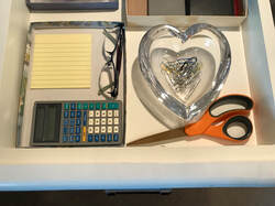 Open drawer (cream colored) with scissors, paperclips in a glass heart shaped container, a cardboard lid with sticky notes, glasses, and a calculator
