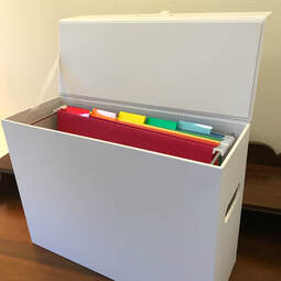 White file box open with rainbow files in order:  red, orange, yellow, green, blue.  The file box has a lid.  The file box is set on a dark wood antique writing desk.  There is a pale yellow wall in the background.