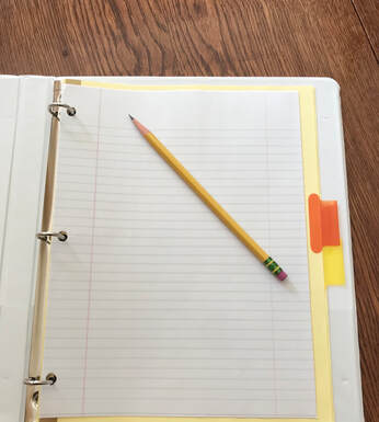 White binder open on a wood desk.  Binder includes lined notebook paper, orange and yellow binder dividers, and a pencil