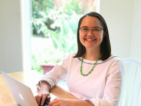 Professional Organizer Laura Sinclair is photographed sitting at a light wood table, with a laptop, wearing a white shirt, green necklace, with a window and greenery in the background