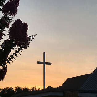 Orange sky behind a cross.  The edges of a flowering tree and church roof are visible.  
