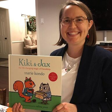 Professional organizer Laura Sinclair holds up Marie Kondo's kids' picture book called Kiki & Jax. Laura is smiling, has shoulder length brown hair, and is wearing a white shirt with a navy sweater.