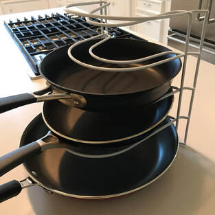 White kitchen counter with edge of stainless steel fridge and black gas stovetop.  Main item is a white five tiered pan organizer rack holding three black colored frying pans with the top two tiers empty.  