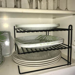 Kitchen cupboard open to show a black three tiered organizing shelf made of metal, each shelf holding a stack of plates.  All plates are white.  Each shelf has 4 to 6 plates, some square and some round of varying sizes.