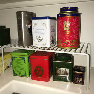 Cream colored pantry shelf holds four tins of tea.  Then, a white plastic shelf organizer creates an additional shelf for three tins of tea.  Tins of tea range from red, white, light green, dark green, silver, and brown, respectively.  