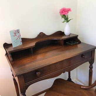 Square photo of dark wood antique writing desk.  The edge of the dark wood chair is visible in the bottom right corner.  The desk is cleared off with two items: a small white smooth vase with pink plastic flowers.  On the left of the desk, there is a greeting card with aqua background and matching pink flowers.  