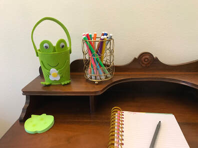 Corner of a brown wooden antique desk with several items for kids: a notebook and pen, green post-it notes shaped like an apple, colorful markers in a decorative gold colored holder, and a frog shaped felt basket