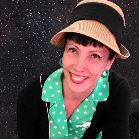 Headshot of Kammy Lee smiling and wearing a turquoise dress with white hearts, a black sweater, green hoop earrings, and a brown hat.