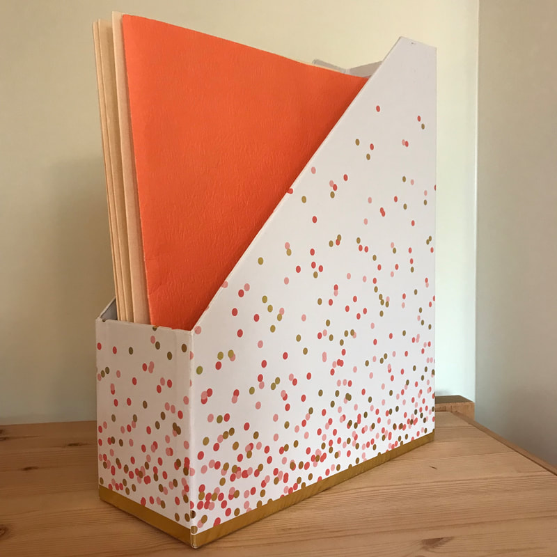 Wooden shelf with white file box (also called a vertical magazine holder).  It is white with a gold trim and pink and gold spots decorating it.  Inside are four tan colored file folders and one orange file folder.  Background is a pale yellow wall.  