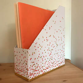 Edge of wooden shelf with a white vertical organizer with pink and orange dots; inside vertically standing up are three folders - one bright orange and two a light tan