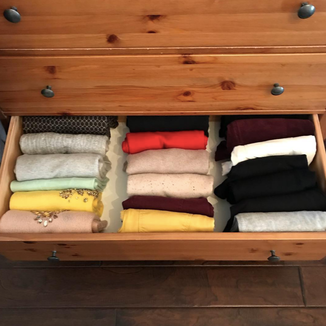 Photo of a wooden dresser with drawers.  One drawer is open to show three rows of vertically folded and organized sweaters, ranging in color from muted colors and greys, tans, and a few dark navy ones.  