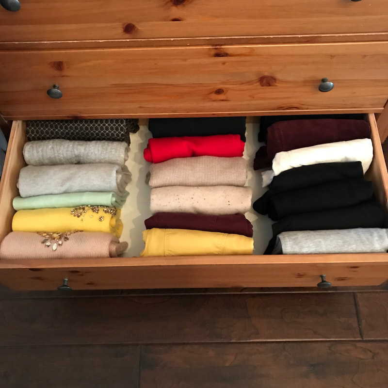 Photo of sweaters folded vertically and stored in a wooden chest of drawers.  The drawer is open, and the sweaters range from greys, tans, yellows, and one red.  